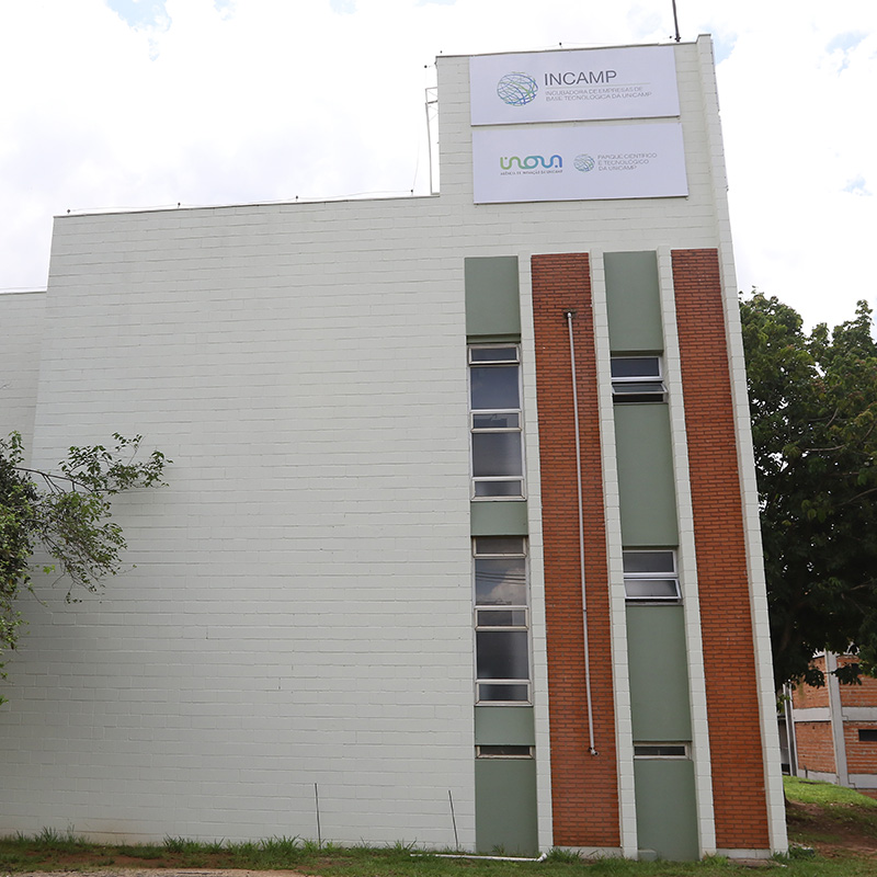 incamp Foto externa do prédio da Incamp. Imagem registra a fachada do local, que também traz uma placa com o logotipo da Incubadora e da Inova Unicamp. O cenário inclui um céu com nuvens brancas e árvores ao fundo. Fim da descrição.